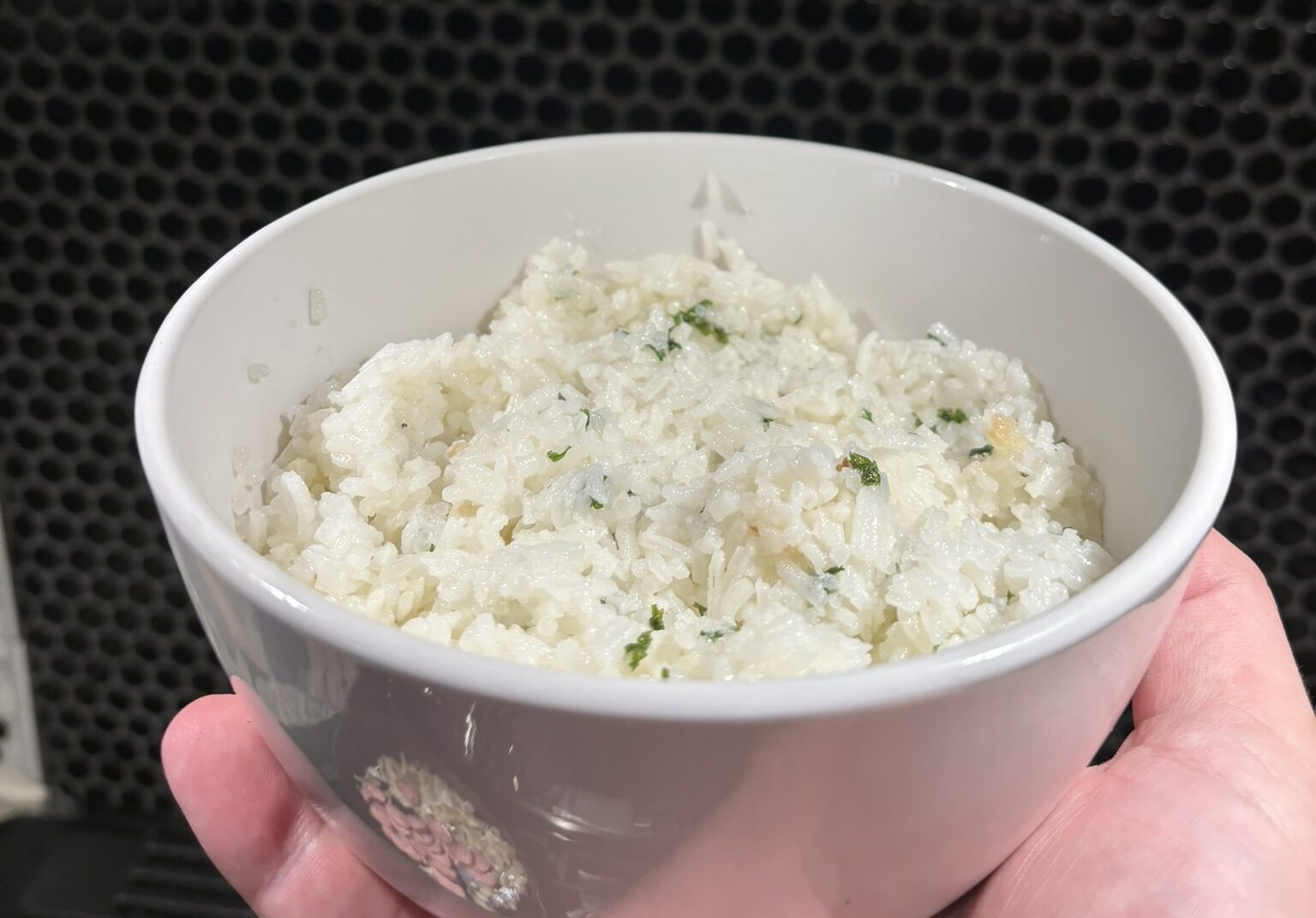 A hand holds a bowl of white rice, with flecks of parsley in it.