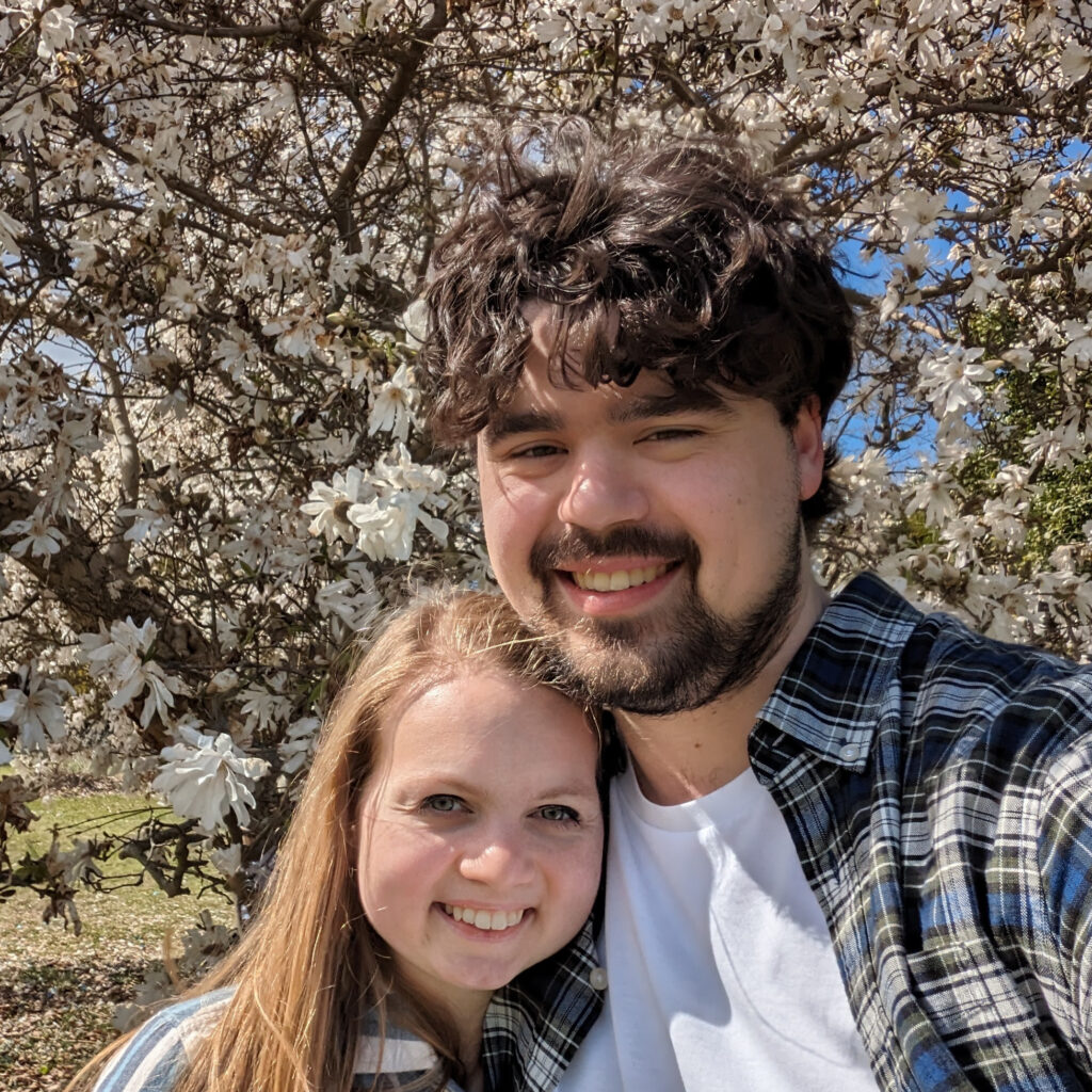 A man and woman, smiling. Behind them is a tree with white flower blossoms.