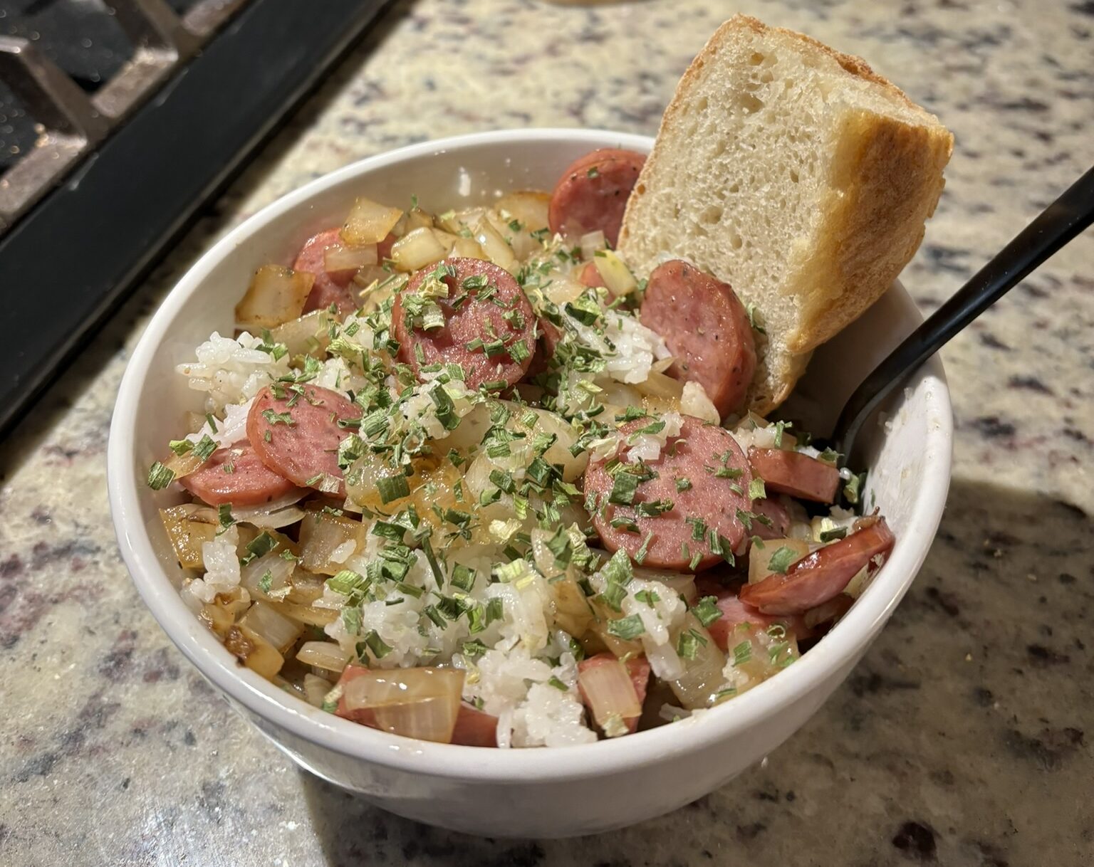 A bowl full of rice, sausage, onions, and a piece of bread sits on a counter top.